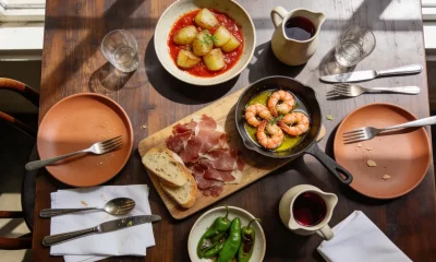 A dark timber restaurant table in natural window light with a board of cured meat and bread, a pan of prawns in oil, a dish of potatoes in red sauce, blistered green peppers and a jug of red wine.