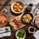 A dark timber restaurant table in natural window light with a board of cured meat and bread, a pan of prawns in oil, a dish of potatoes in red sauce, blistered green peppers and a jug of red wine.