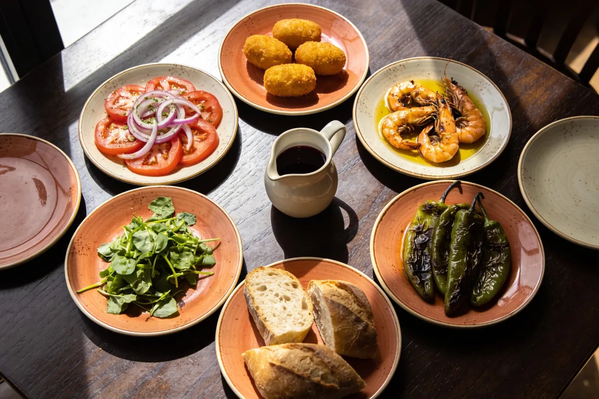 A spread of small plates on a dark timber restaurant table seen from above, including croquetas, a tomato salad, prawns in oil, blistered green peppers, greens and crusty bread beside a jug of red wine.