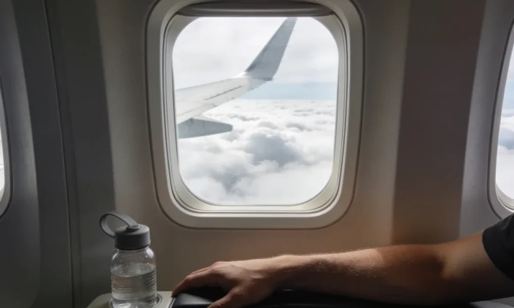 View from an aeroplane window seat with a wing passing over clouds and a person's lower arm resting on the armrest beside a small reusable water bottle in soft daylight.
