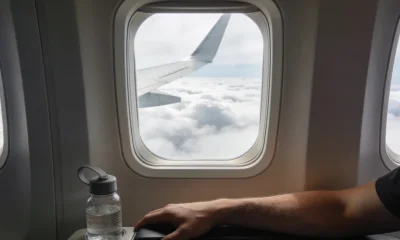 View from an aeroplane window seat with a wing passing over clouds and a person's lower arm resting on the armrest beside a small reusable water bottle in soft daylight.