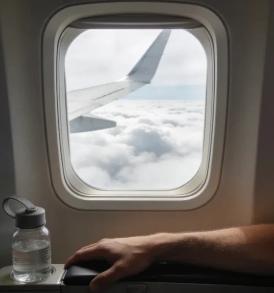 View from an aeroplane window seat with a wing passing over clouds and a person's lower arm resting on the armrest beside a small reusable water bottle in soft daylight.