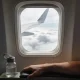 View from an aeroplane window seat with a wing passing over clouds and a person's lower arm resting on the armrest beside a small reusable water bottle in soft daylight.