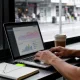 Small-business owner working on a marketing plan at a cafe window table with a laptop and notebook.