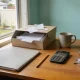 Home-office desk in morning light with an open box of paper receipts, a closed folder, a pocket calculator, a pencil, reading glasses and a mug, beside a window onto a suburban backyard