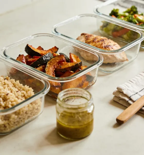 Several unlabelled clear glass containers of prepared meal-prep food, cooked grains, roasted vegetables, a cooked protein and a jar of dressing, in a neat row on a bright kitchen bench with a tea towel and a wooden spoon.