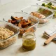 Several unlabelled clear glass containers of prepared meal-prep food, cooked grains, roasted vegetables, a cooked protein and a jar of dressing, in a neat row on a bright kitchen bench with a tea towel and a wooden spoon.