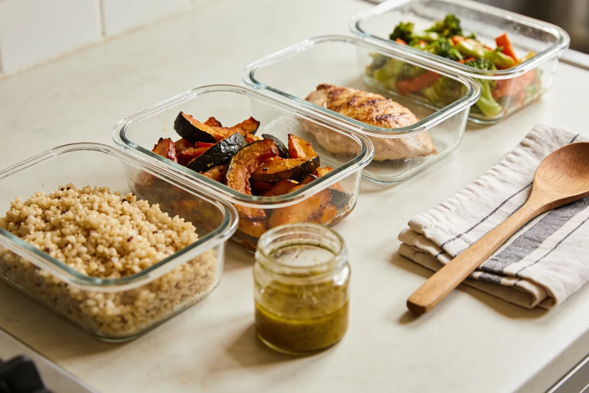 Several unlabelled clear glass containers of prepared meal-prep food, cooked grains, roasted vegetables, a cooked protein and a jar of dressing, in a neat row on a bright kitchen bench with a tea towel and a wooden spoon.
