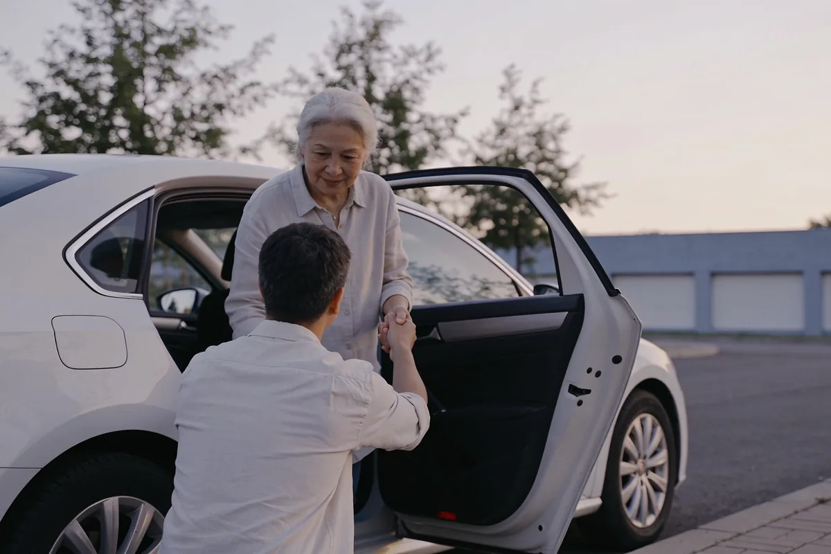 A driver helping an older passenger out of a car outside a medical centre in the morning.