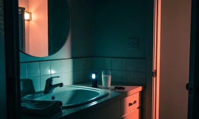 Dimly lit Australian bathroom seen from the doorway at night, with a glass of milk, a small lit torch and a car key on the vanity beside a round mirror and folded towel, warm light spilling in from the hall