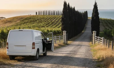Plain white van parked on a gravel vineyard driveway with a door open, a windbreak of cypress trees and vine rows rising behind it and a soft glimpse of the bay beyond