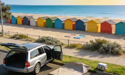 Silver station wagon parked at the kerb above a bayside beach, tailgate open, with a folded beach tent and a cooler bag on the grass verge and a long row of brightly coloured bathing boxes on the sand beyond