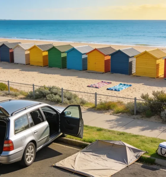 Silver station wagon parked at the kerb above a bayside beach, tailgate open, with a folded beach tent and a cooler bag on the grass verge and a long row of brightly coloured bathing boxes on the sand beyond