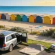 Silver station wagon parked at the kerb above a bayside beach, tailgate open, with a folded beach tent and a cooler bag on the grass verge and a long row of brightly coloured bathing boxes on the sand beyond