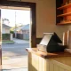 Cafe counter in early morning light with a closed till, a stack of plain takeaway cups, a folded cloth and an apron on a hook, opening onto a quiet suburban street through the doorway