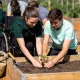 A support worker guiding a young adult's hands as they plant a seedling in a raised garden bed at a sunny Melbourne community garden.