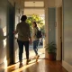 A support worker holding the front door open as a young woman with a backpack steps out of a sunlit Melbourne terrace house.