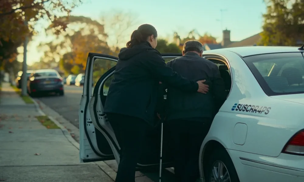 A support worker helping a man with a walking cane into an unmarked white car on a quiet suburban Melbourne street in early morning light.