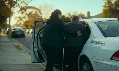 A support worker helping a man with a walking cane into an unmarked white car on a quiet suburban Melbourne street in early morning light.