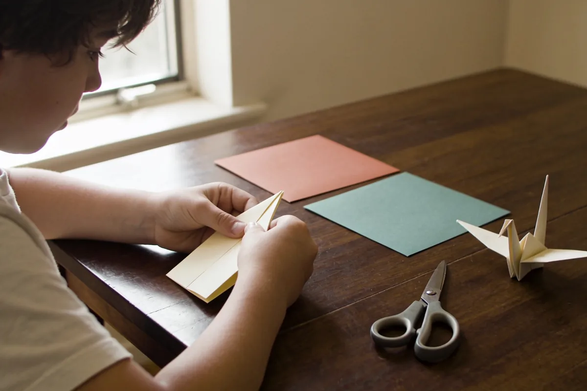 child folding origami with scissors and paper at a wooden table