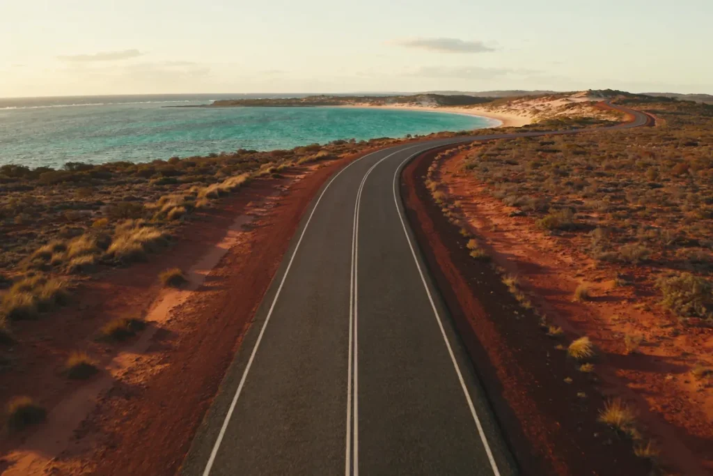 Elevated view of a long empty coastal road on Western Australia's Coral Coast bending toward a turquoise bay on the horizon, dry red earth and low scrub on either side in late-afternoon light