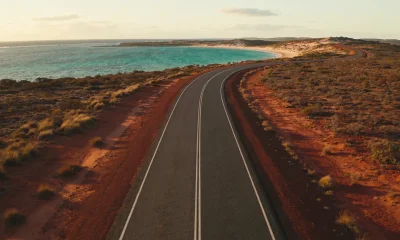 Elevated view of a long empty coastal road on Western Australia's Coral Coast bending toward a turquoise bay on the horizon, dry red earth and low scrub on either side in late-afternoon light