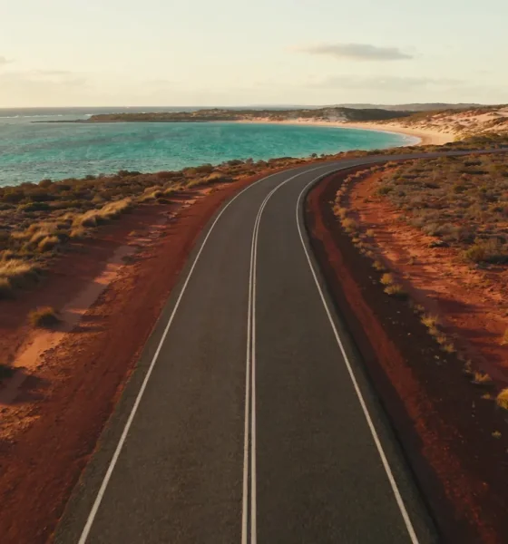 Elevated view of a long empty coastal road on Western Australia's Coral Coast bending toward a turquoise bay on the horizon, dry red earth and low scrub on either side in late-afternoon light