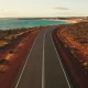 Elevated view of a long empty coastal road on Western Australia's Coral Coast bending toward a turquoise bay on the horizon, dry red earth and low scrub on either side in late-afternoon light