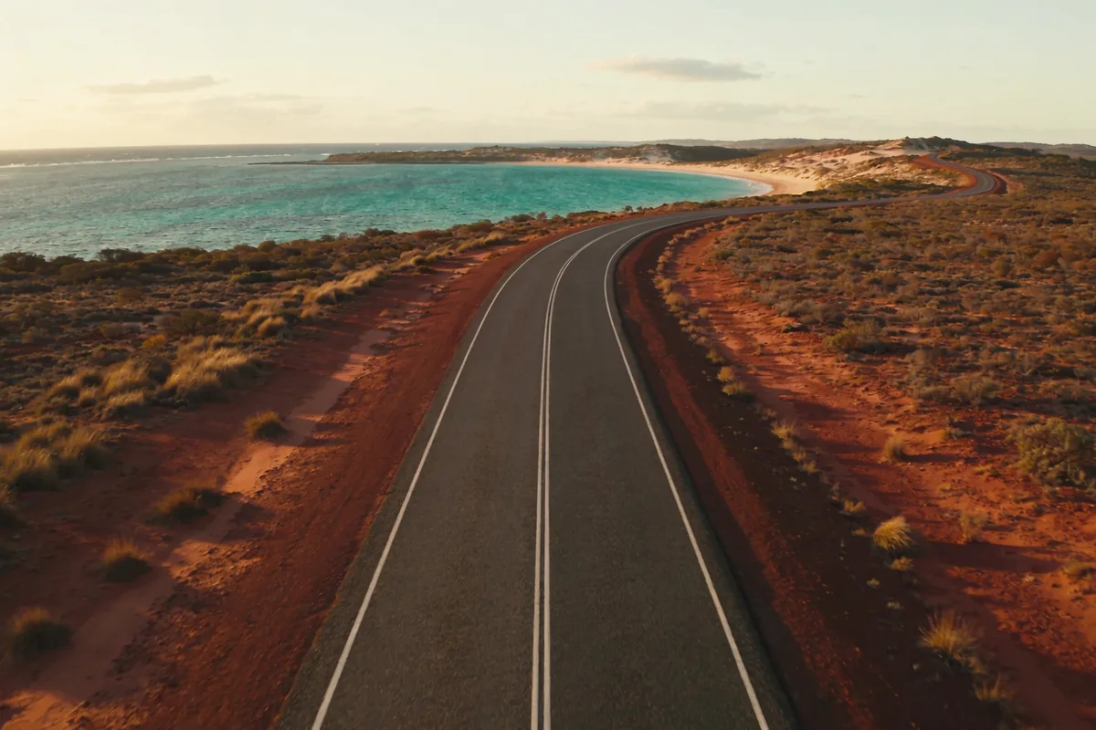 Elevated view of a long empty coastal road on Western Australia's Coral Coast bending toward a turquoise bay on the horizon, dry red earth and low scrub on either side in late-afternoon light