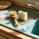 A pale natural dish soap bar and a wooden dish brush on a draining rack beside a ceramic kitchen sink in warm afternoon light.