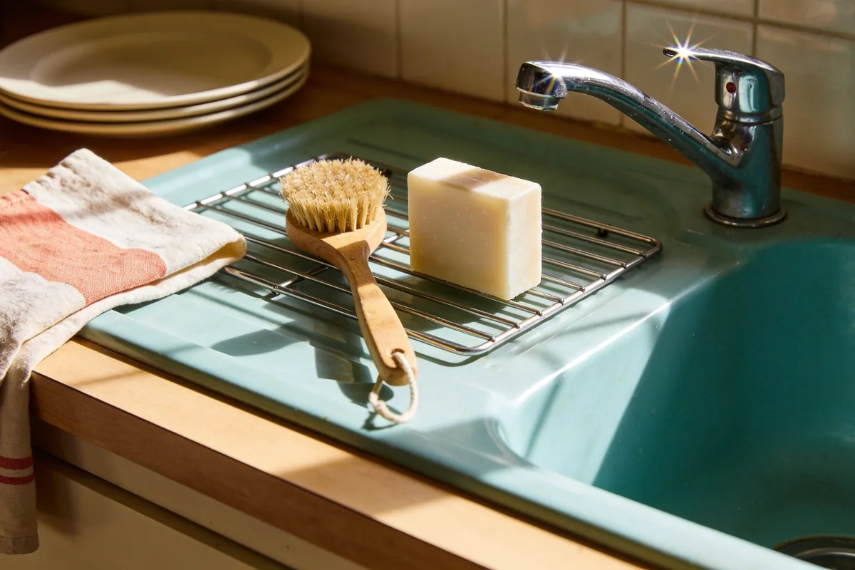 A pale natural dish soap bar and a wooden dish brush on a draining rack beside a ceramic kitchen sink in warm afternoon light.