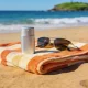An unlabelled metal sunscreen tin and sunglasses resting on a striped towel on an Australian beach with ocean and headland behind.