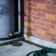 Outdoor storage hot water cylinder on a concrete pad against a red brick wall on a suburban side path, with insulated pipework, a brass valve at the base, a damp patch on the concrete and a coiled green hose beside a galvanised bucket