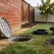 Open plastic drain inspection point set in a backyard lawn beside a red brick wall, its lid resting on the grass with a coil of camera cable running from a reel on the turf beside a damp patch