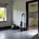 Empty living room mid-clean in bright morning light, with bare floorboards carrying faint scuff marks, a vacuum cleaner against the skirting, a bucket with a cloth draped over the rim, cleaning spray and paper towel by the window, and a bare kitchen through the doorway