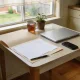 A small kitchen table by a curtained window in an Australian home in mid-morning light, with an open manila folder and blank pages, a closed silver laptop, a phone and a mug of tea beside a potted plant, and a garden beyond the glass