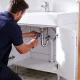 A plumber tightening pipework under a bathroom vanity in a bright Australian home.