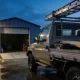 Silver work ute with a ladder rack, toolbox and timber in its tray, parked on a wet concrete apron in front of a corrugated iron workshop with an open roller door in the early morning