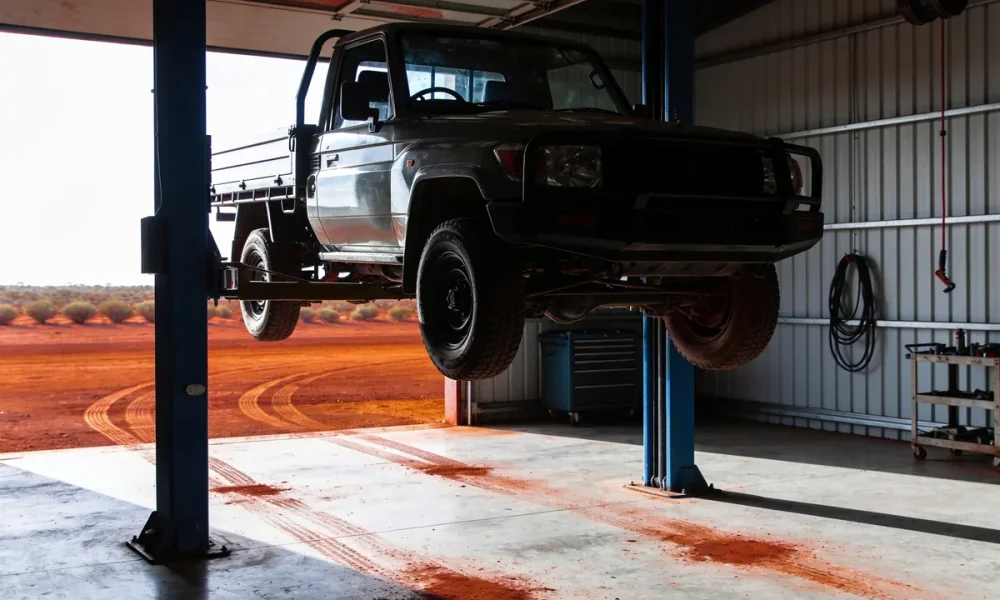 Ute raised on a two-post hoist inside a regional workshop, red dust across the concrete floor, with red dirt and low scrub visible through the open roller door