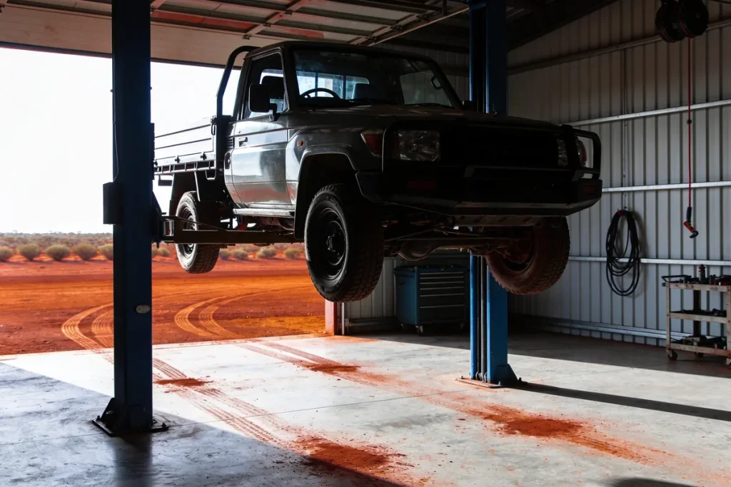 Ute raised on a two-post hoist inside a regional workshop, red dust across the concrete floor, with red dirt and low scrub visible through the open roller door