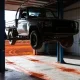 Ute raised on a two-post hoist inside a regional workshop, red dust across the concrete floor, with red dirt and low scrub visible through the open roller door