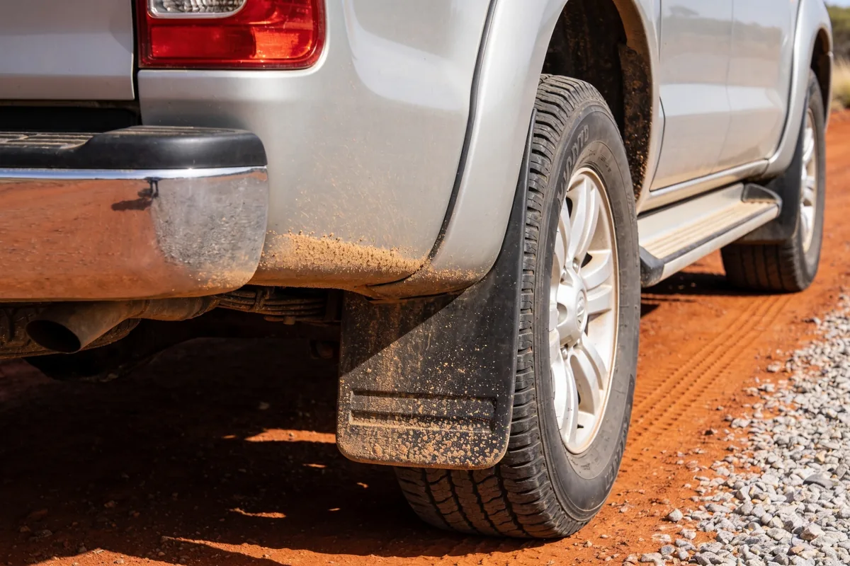Lower rear quarter of a dusty ute on a red dirt road, with dust along the wheel arch and mudflap and gravel scattered beside the rear wheel