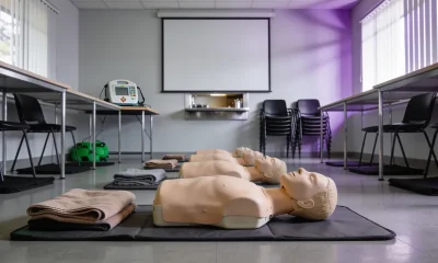 Community training room set up for a first aid course, with a row of CPR manikins on floor mats, folded blankets and a training defibrillator on a table below a blank projector screen