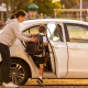 A parent helping a child with a backpack into the back seat of a waiting car in a school pickup zone in the late afternoon.