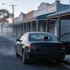 A plain dark sedan parked at the kerb of a country town main street at first light, its rear door open with a soft travel bag standing on the footpath beside it, empty veranda-fronted shopfronts running down the street and mist over the paddocks at the far end