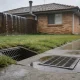 A wet Australian backyard in the rain, with a stormwater grate set into the concrete path and a grated pit in the grass beside it, and an older brick house and timber fence behind.