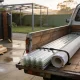 Flatbed ute loaded with translucent corrugated roofing sheets and a wrapped roll of flashing strapped to its timber tray, with lengths of timber stacked on the ground beside an open gate and a pergola frame in the yard beyond