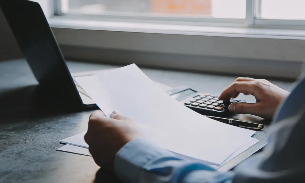 Over-shoulder view of a person's hands reviewing blank papers beside a calculator and a pen at a sunlit desk in a small Australian business office, soft morning light, no readable text.