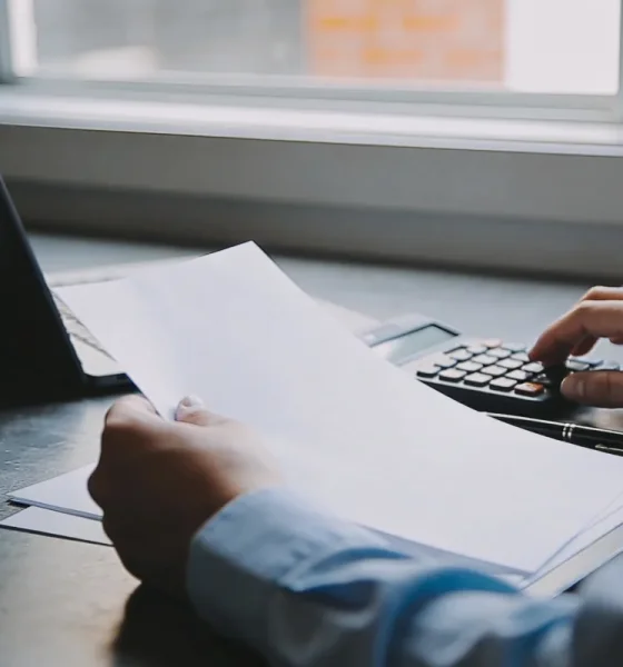 Over-shoulder view of a person's hands reviewing blank papers beside a calculator and a pen at a sunlit desk in a small Australian business office, soft morning light, no readable text.