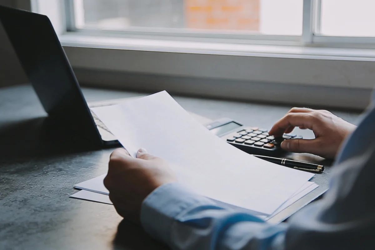 Over-shoulder view of a person's hands reviewing blank papers beside a calculator and a pen at a sunlit desk in a small Australian business office, soft morning light, no readable text.
